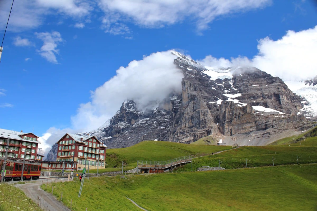 Paysage alpin au glacier de l’Eiger avec un plateau verdoyant, un hôtel de montagne et des parois rocheuses abruptes sous un ciel bleu.