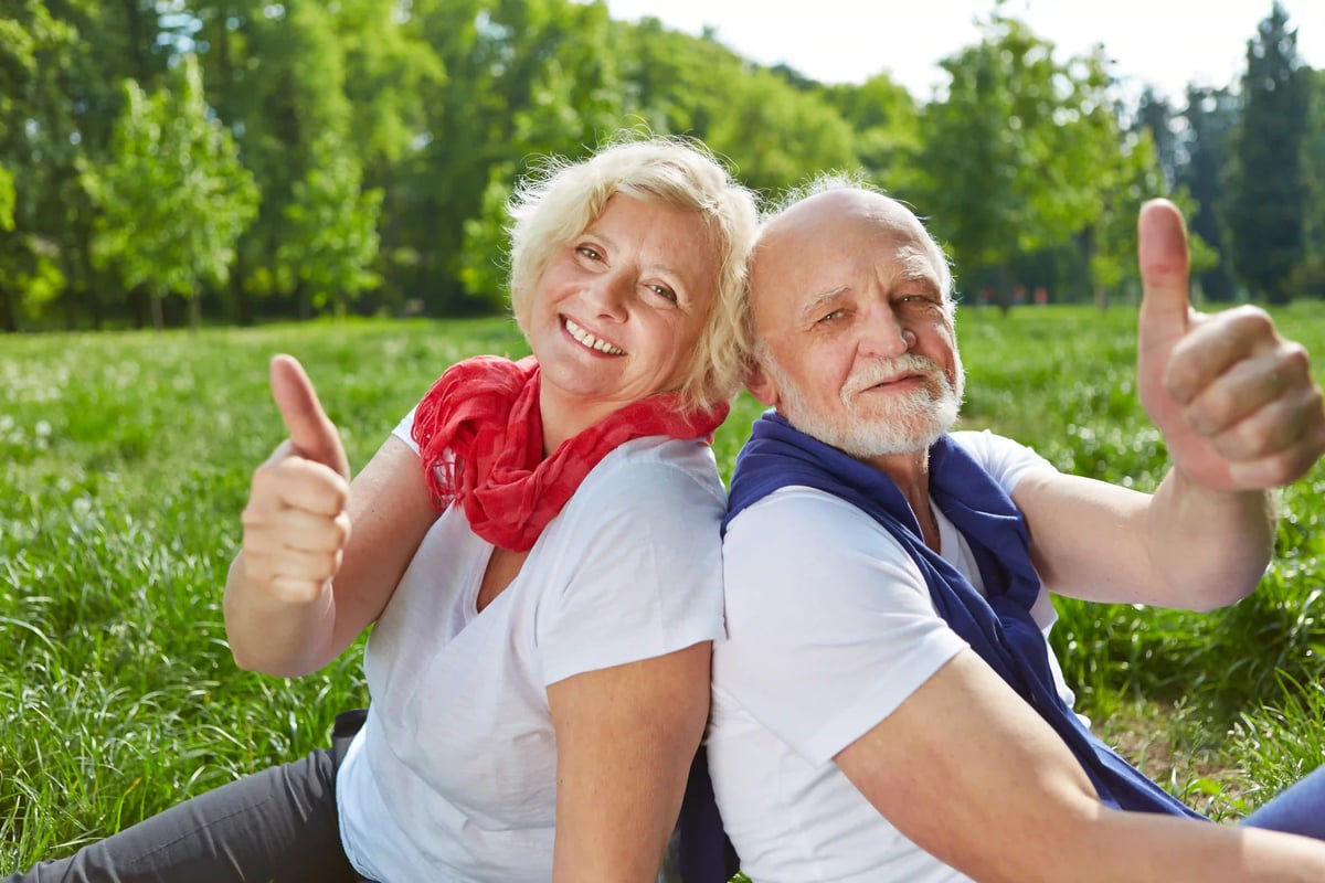 Couple senior assis dans la verdure, souriant à la caméra et montrant un geste de satisfaction avec le pouce levé.