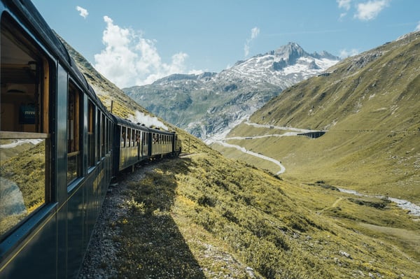 Historischer Zug fährt durch ein alpines Tal mit grünen Hängen und schroffen Bergen, aufgenommen aus einem Waggon