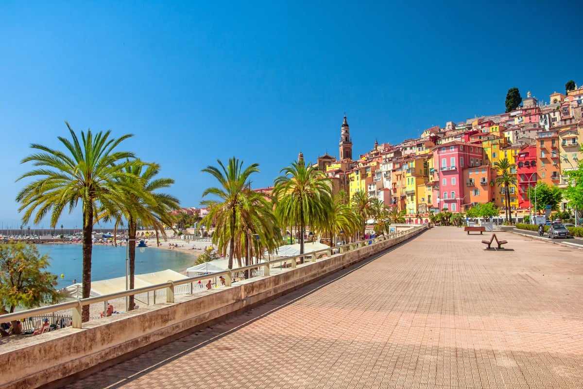 Maisons colorées de Menton le long de la promenade en bord de mer au coucher du soleil, reflets sur la mer calme et palmiers sous un ciel aux teintes pastel