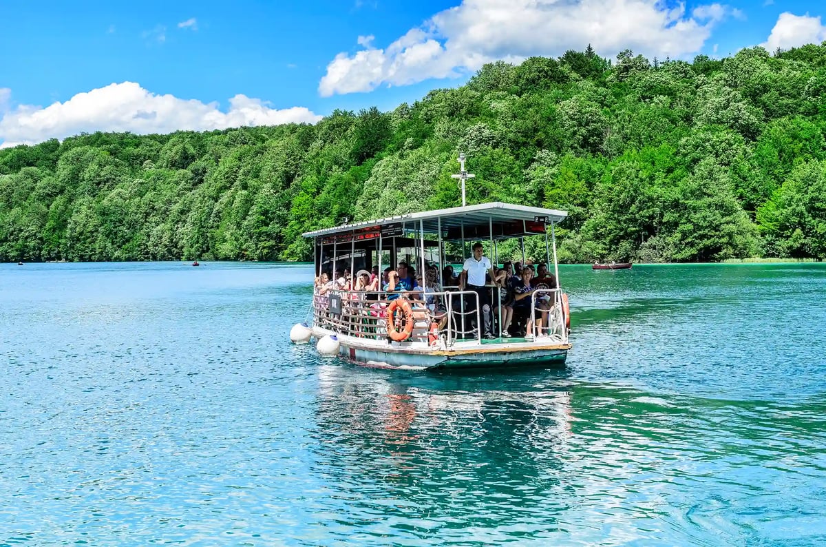Ausflugsboot mit Besuchern auf dem türkisfarbenen Kozjak-See, umgeben von bewaldeten Hügeln und blauem Himmel