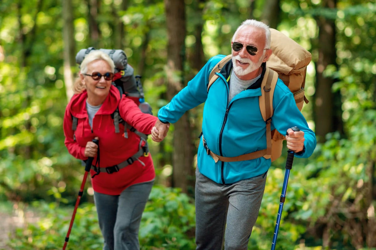 Älteres Wanderpaar mit Rucksäcken und Wanderstöcken geht durch einen sonnigen Wald und lächelt