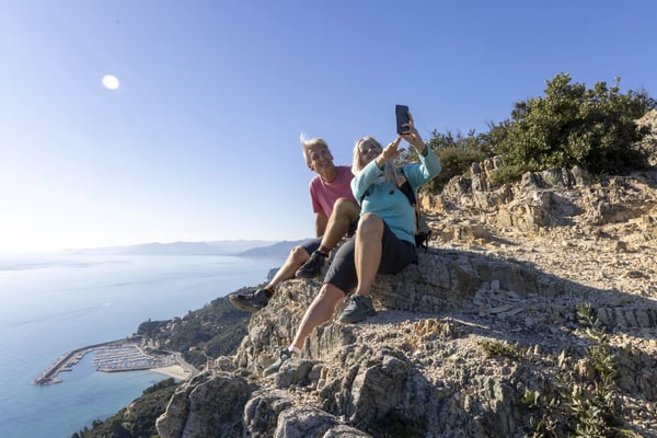 Wanderndes Paar sitzt auf einem Felsen mit Blick auf Meer, Küste und Hafen in Ligurien