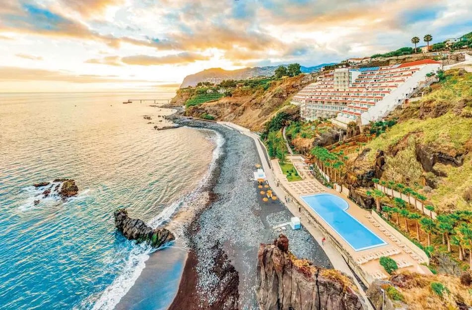 Terrassenförmig angelegte Hotelanlage an felsiger Küste mit Meerblick, Küstenweg, Poolbereich und Sonnenuntergang am Horizont.