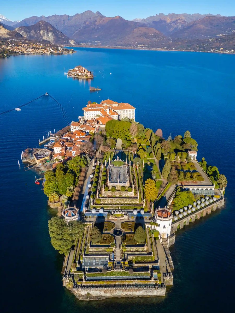 Vue aérienne d’une île avec palais et jardins symétriques sur un lac bleu profond, montagnes à l’horizon.