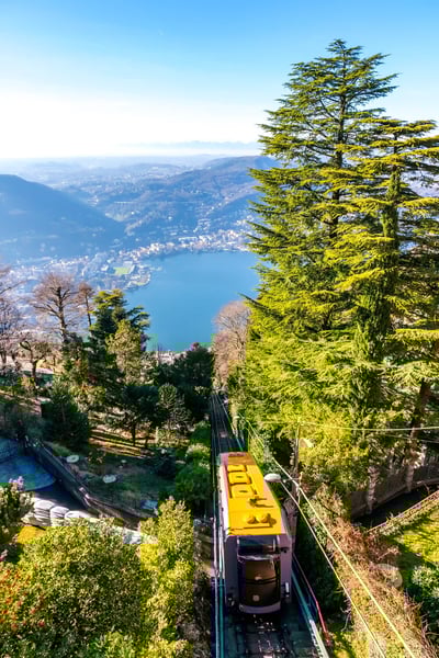 Funiculaire jaune descend à travers une forêt dense, avec un lac bleu et une ville au loin.