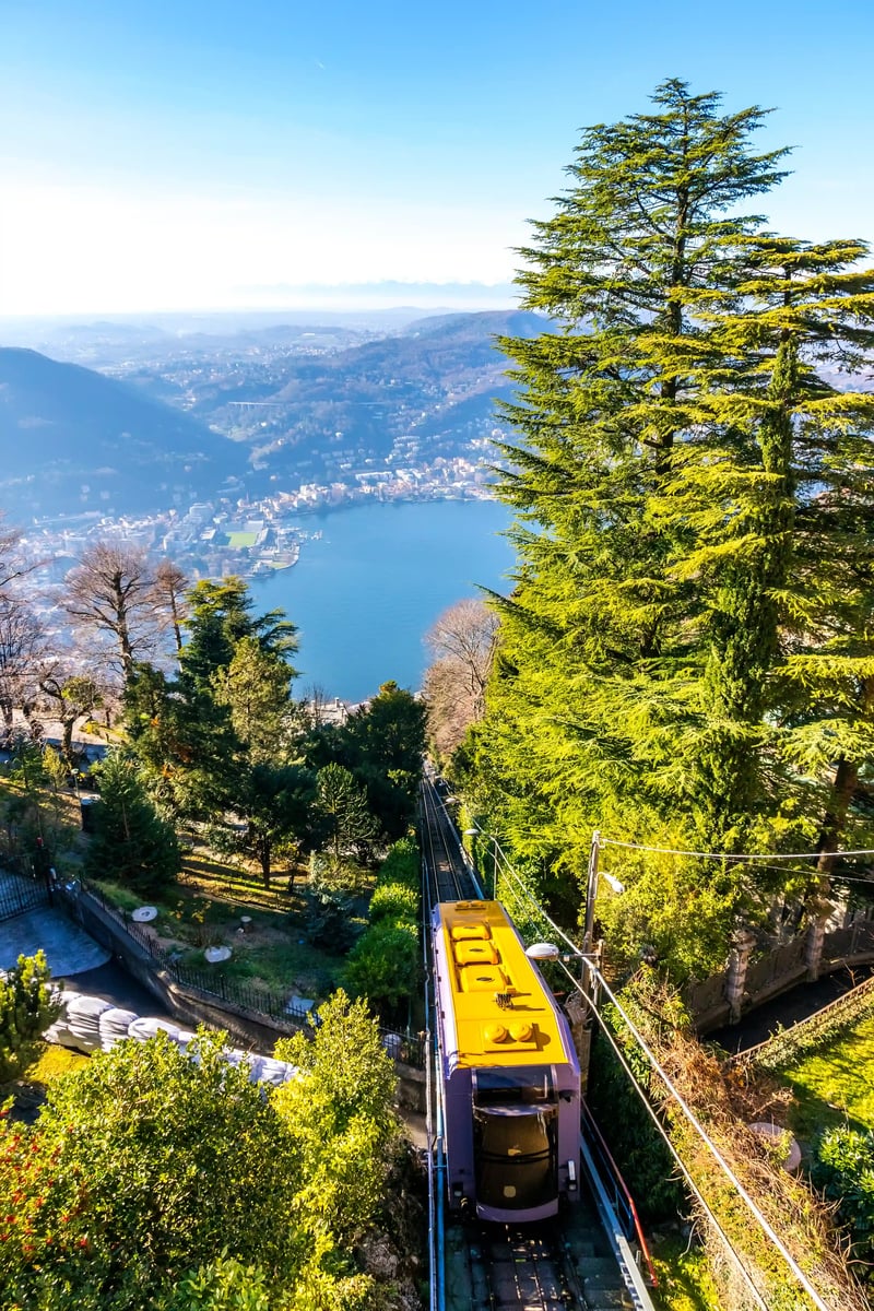 Funiculaire jaune descend à travers une forêt dense, avec un lac bleu et une ville au loin.