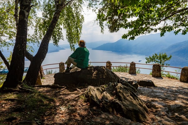 Personne assise sur un rocher sous les arbres, face à un vaste lac entre des montagnes brumeuses.