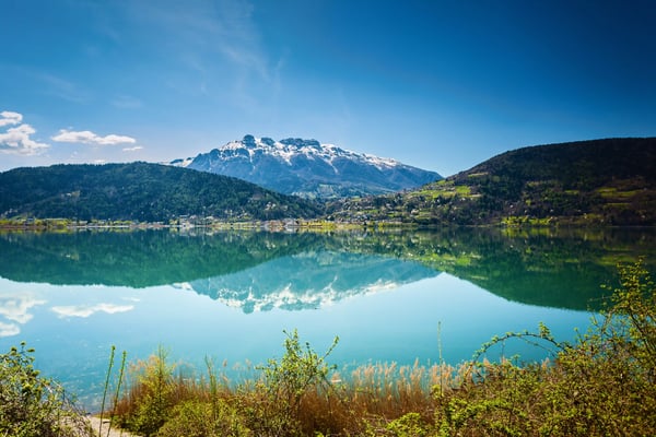 Ruhiger Bergsee mit Spiegelung schneebedeckter Gipfel und grünen Hügeln unter blauem Himmel