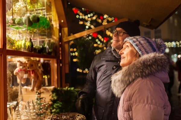 Älteres Paar mit Geschenken auf Weihnachtsmarkt vor beleuchtetem Baum