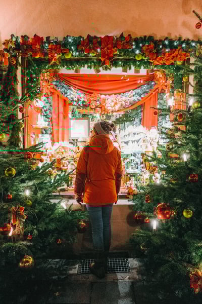 Person in roter Jacke steht vor festlich geschmücktem Weihnachtsmarktstand mit Lichtern und Dekoration