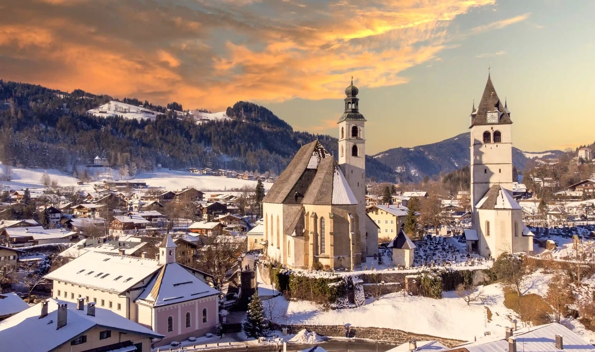 Winterliches Kitzbühel mit Pfarrkirche, schneebedeckten Dächern und Alpenpanorama bei Sonnenuntergang.