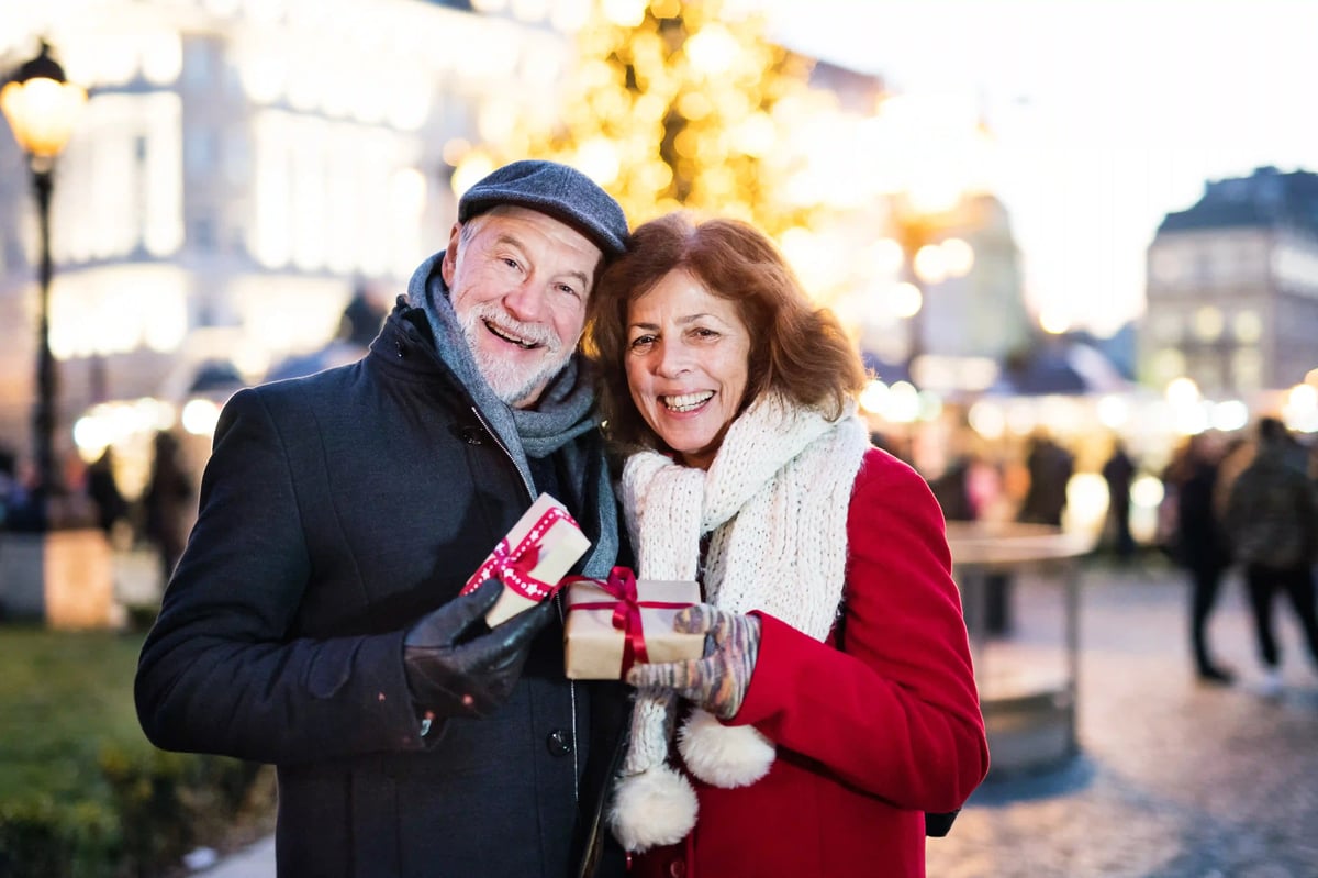 Älteres Paar mit Geschenken auf einem Weihnachtsmarkt vor beleuchtetem Baum