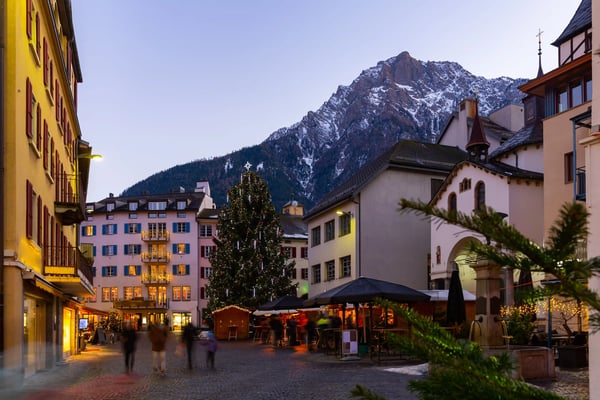 Dorfplatz in den Alpen im Winter mit beleuchtetem Weihnachtsbaum und verschneiten Bergen im Hintergrund