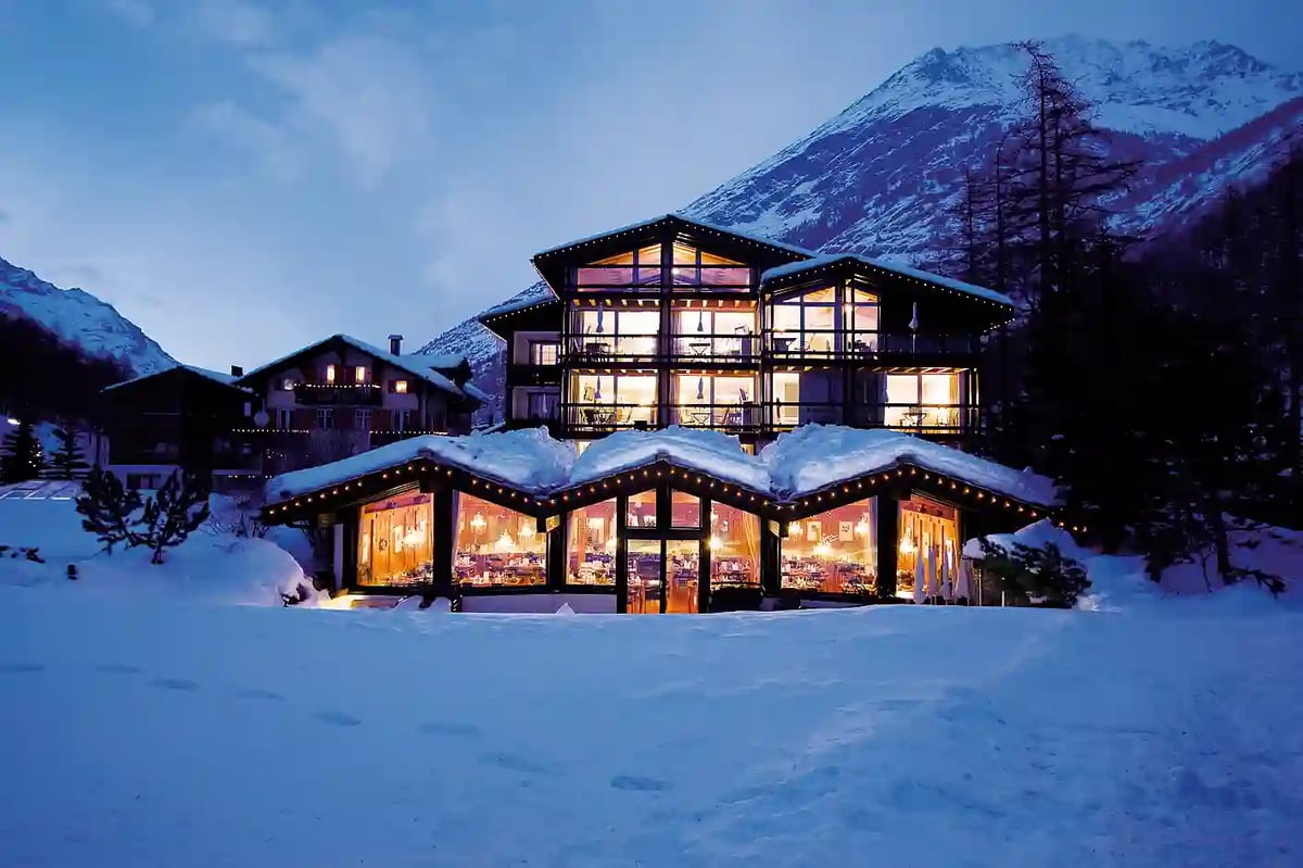 Modernes Hotel in verschneiter Berglandschaft mit beleuchteten Innenräumen bei Dämmerung