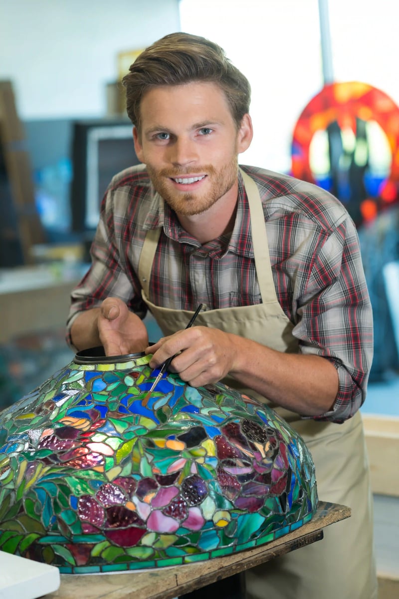 Artisan dans un atelier peignant une lampe en verre multicolore, penché sur le motif en mosaïque.