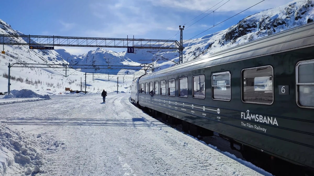 alt="Ein dunkelgrüner Zug mit der Aufschrift Flåmsbana steht an einer verschneiten Bahnstation in den Bergen, während eine Person auf den Gleisen geht.