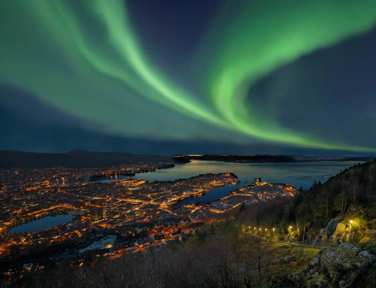 Grüne Polarlichter erstrecken sich am nächtlichen Himmel über der hell erleuchteten Stadt Bergen an einer Meeresbucht, fotografiert von einem Berghang.