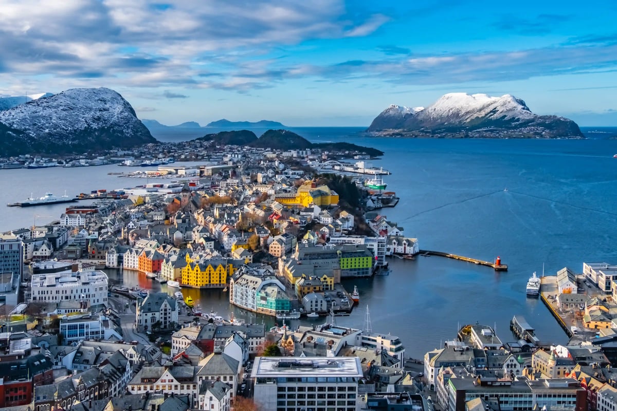 Blick von oben auf die farbenfrohe Stadt Ålesund, die sich auf Inseln im blauen Meer erstreckt, umgeben von schneebedeckten Bergen unter bewölktem Himmel.