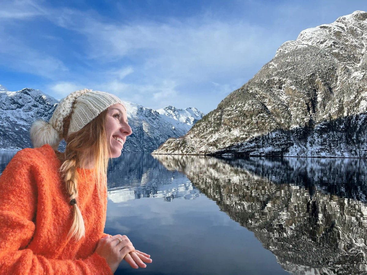 Eine lächelnde Frau mit orangerotem Pullover und weisser Mütze blickt auf den spiegelglatten Geirangerfjord, in dem sich die verschneiten Berge reflektieren.