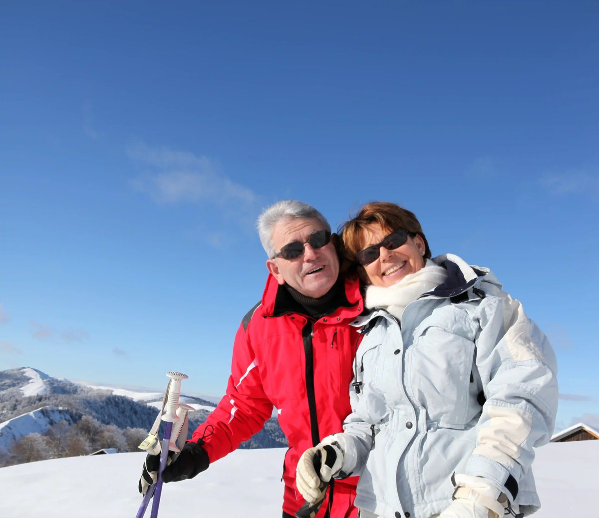 Lächelndes Paar in Winterkleidung mit Skistöcken vor verschneiter Berglandschaft