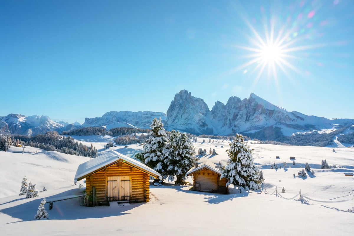 Verschneite Seiser Alm mit Holzhütten, Tannen und Dolomitengipfeln bei strahlendem Sonnenschein