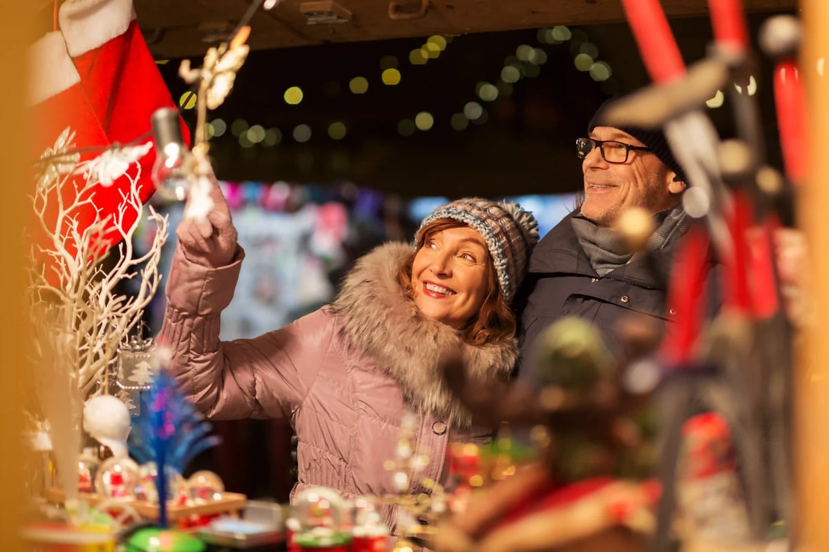 Couple en vêtements d’hiver souriant devant un stand de marché de Noël décoré de guirlandes lumineuses