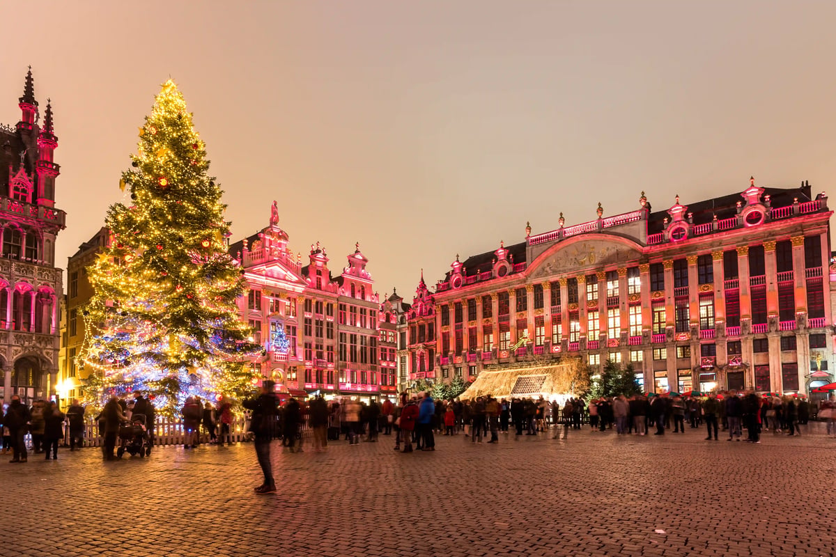 Grand sapin de Noël illuminé sur la Grand-Place de Bruxelles, entouré de bâtiments historiques décorés et de visiteurs profitant des festivités de l’Avent.