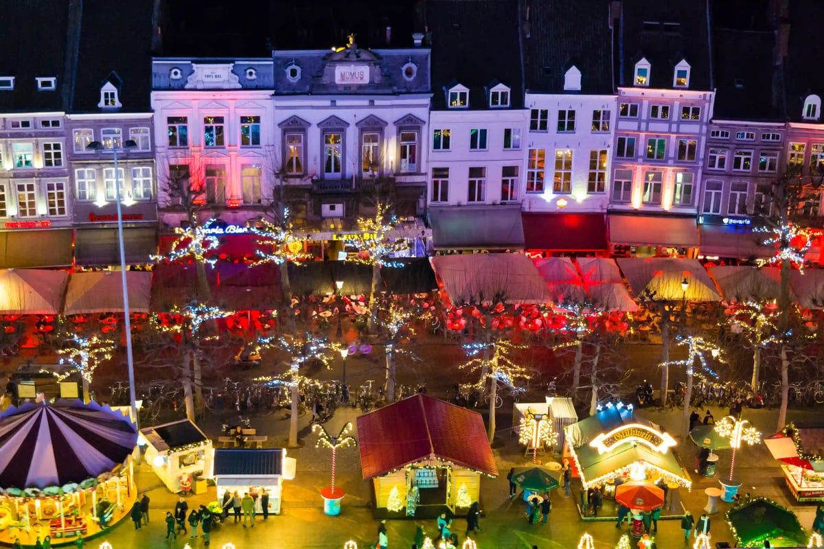 Vue aérienne d’un marché de Noël à Maastricht avec chalets illuminés, décorations festives et visiteurs se promenant dans une ambiance hivernale chaleureuse.