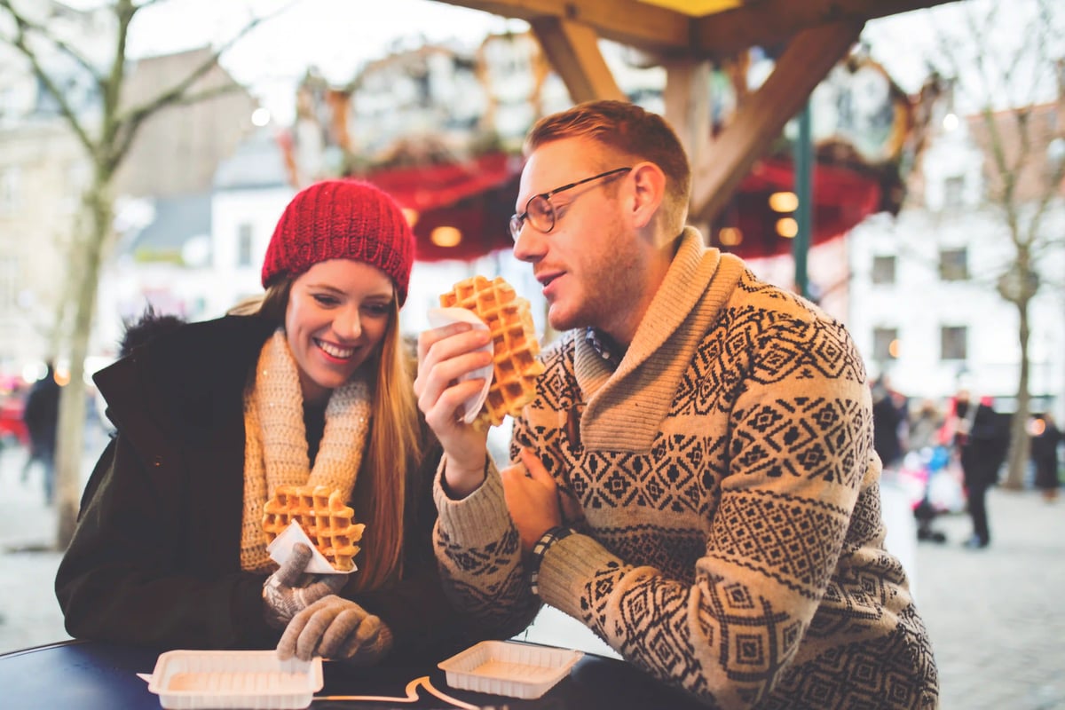 Deux voyageurs dégustent des gaufres sur un marché de Noël, vêtus de vêtements d’hiver et profitant d’une atmosphère conviviale et festive.