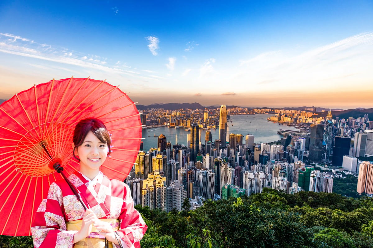 Eine lächelnde Frau im rot-weissen Kimono mit rotem Papierschirm vor der weiten Skyline von Hongkong und dem Victoria Harbour bei Sonnenuntergang.