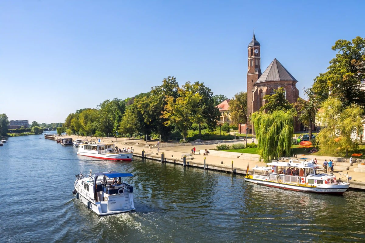 Flussufer in Brandenburg an der Havel mit Ausflugsschiffen, Promenade und Kirche zwischen Bäumen