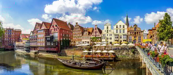 Historischer Hafen in Lüneburg mit altem Holzkrahn am Ufer, Boot im ruhigen Fluss und Cafés unter Sonnenschirmen.