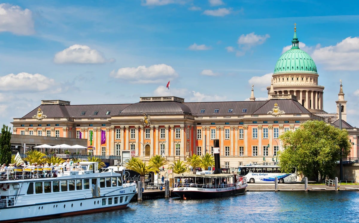 Uferfront in Potsdam mit klassischem Gebäude und grüner Kuppel, davor Schiffe auf dem Fluss bei sonnigem Wetter