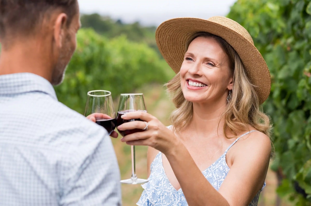 Frau mit Strohhut stoesst mit einem Mann im Weinberg mit Rotwein an.