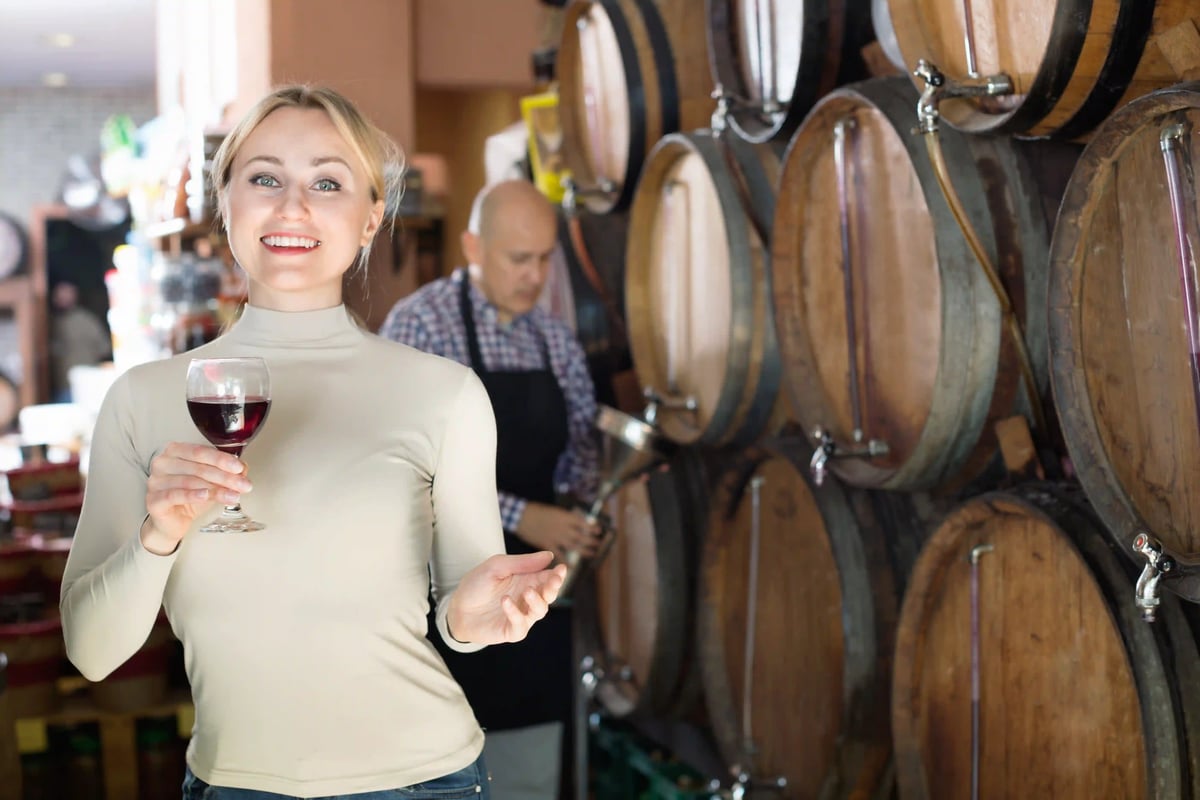Femme tenant un verre de vin rouge dans une cave avec fûts en bois, vigneron travaillant à l arrière-plan