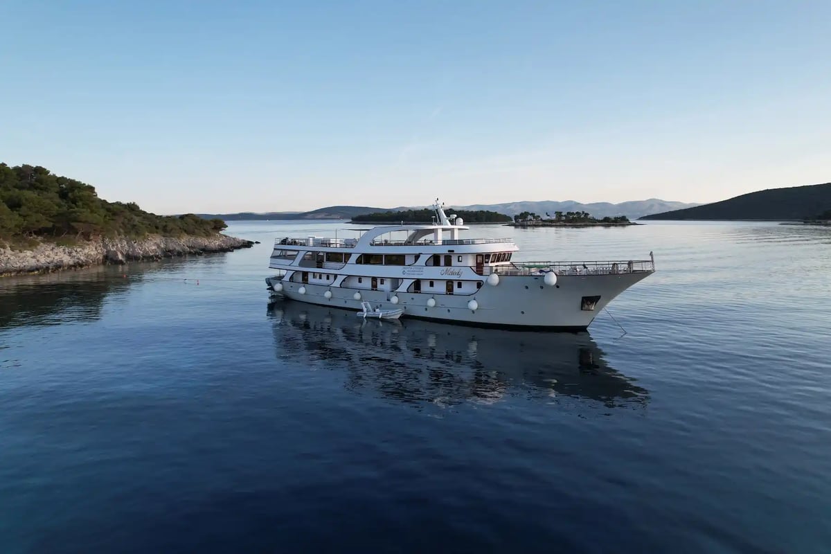 Aussenansicht eines weissen Kreuzfahrtschiffes auf ruhigem Meer vor bewaldeter Küste