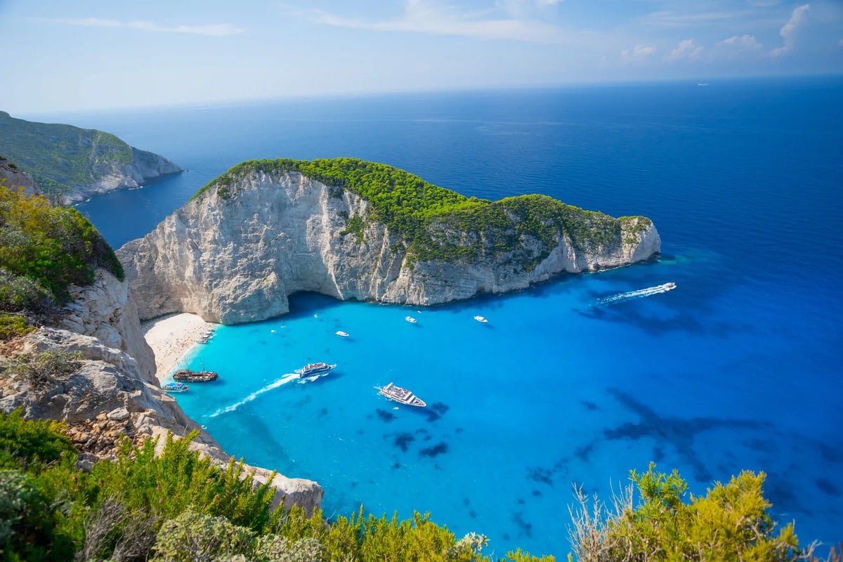 Vue depuis une falaise sur la célèbre baie de Navagio à Zakynthos avec une plage de sable fin, des parois rocheuses abruptes et plusieurs bateaux dans une eau de mer limpide et d'un bleu turquoise intense.