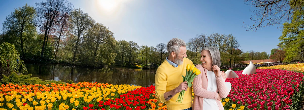 Ein älteres, lächelndes Paar steht inmitten von weitläufigen, farbenfrohen Tulpenfeldern in Gelb, Orange und Rot. Der Mann im gelben Pullover hält einen Strauss gelbe Tulpen und blickt die Frau im rosa Cardigan glücklich an; im Hintergrund ist ein Teich zu sehen.