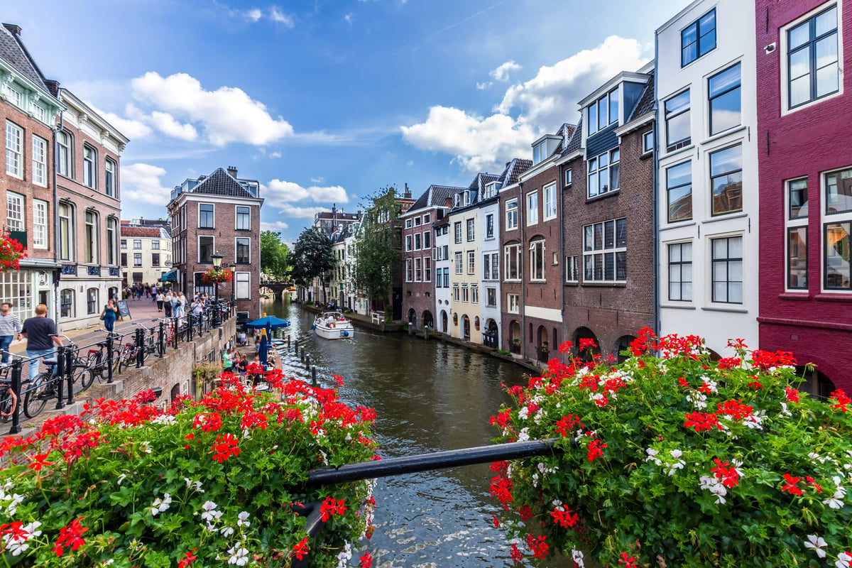 Blick auf einen von historischen, mehrstöckigen Stadthäusern gesäumten Kanal in Utrecht, auf dem ein kleines Boot fährt. Im Vordergrund hängen üppige, rot-weiss blühende Geranien an einem Geländer.