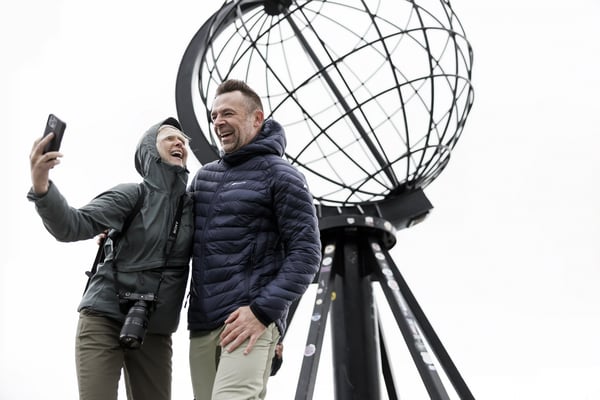 Reisepaar macht ein Foto vor dem Nordkap Globus Denkmal bei bewölktem Himmel