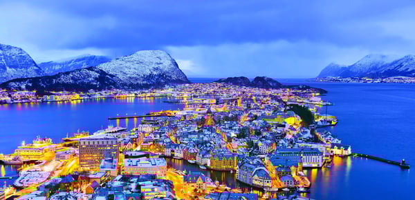 Luftaufnahme der Stadt Ålesund im Winter mit beleuchteten Strassen, Hafen und schneebedeckten Bergen