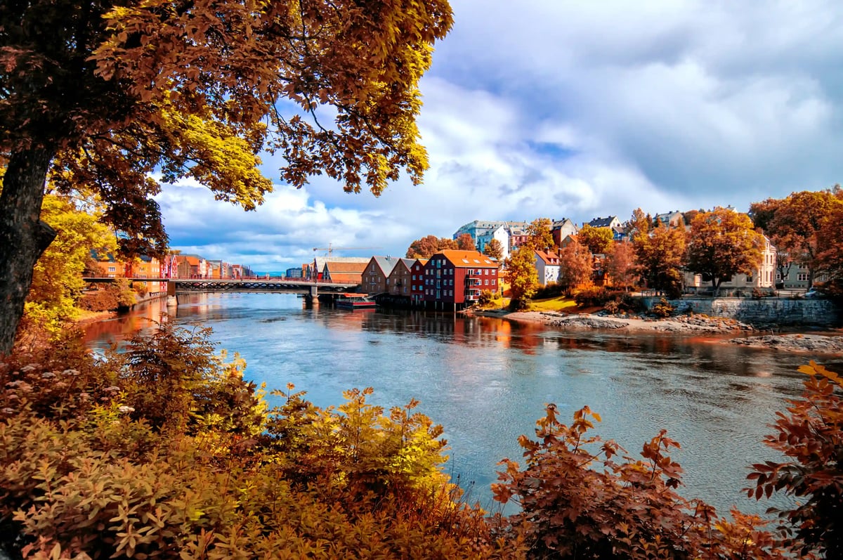 Vue sur une rivière à Trondheim sur les entrepôts historiques colorés au bord de l'eau sous une lumière automnale avec des feuilles colorées sur les arbres au premier plan.