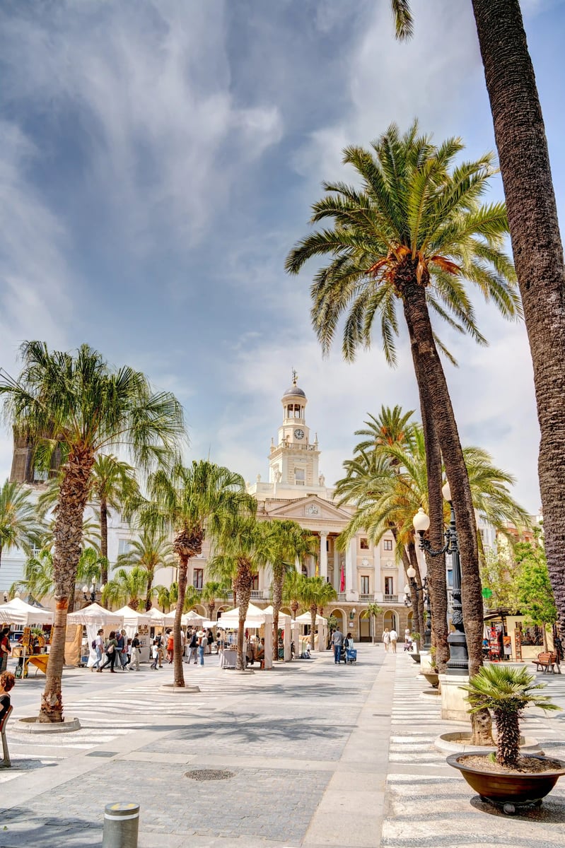 Belebter Platz in Cádiz mit barocker Kathedrale, Palmen und zahlreichen Menschen bei sonnigem Wetter