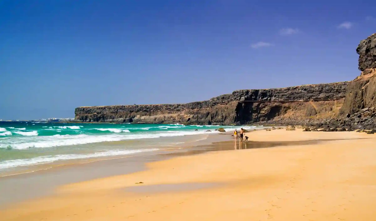Natürlicher Sandstrand mit Felsklippen, türkisfarbenem Wasser und Menschen beim Spazieren am Ufer
