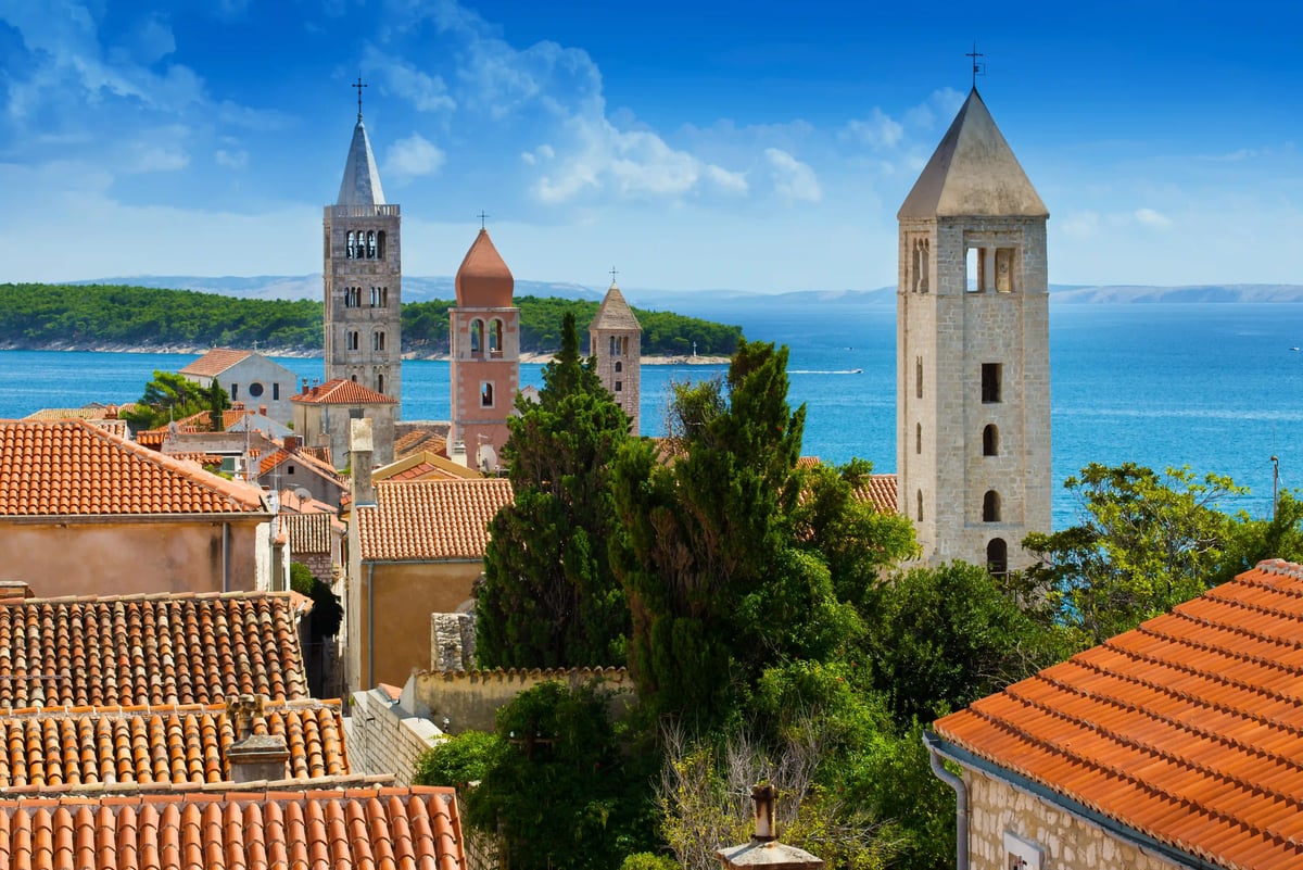 Vue sur la vieille ville de l’île de Rab avec des toits rouges, des clochers et la mer bleue en arrière-plan.