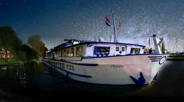 Flusskreuzfahrtschiff MS Olympia bei Nacht auf der Maas vor beleuchtetem Ufer
