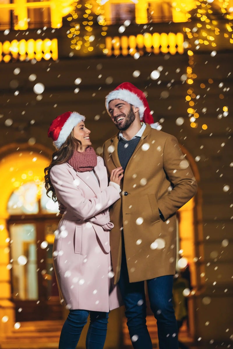Couple souriant portant manteaux d hiver et bonnets de Noël sous la neige devant un bâtiment illuminé