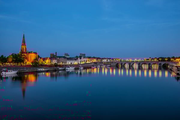 Abendliche Ansicht der Maas mit historischer Brücke, Stadtlichtern und Spiegelungen im Wasser