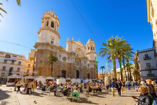 Belebter Platz in Cádiz mit barocker Kathedrale, Palmen und zahlreichen Menschen bei sonnigem Wetter